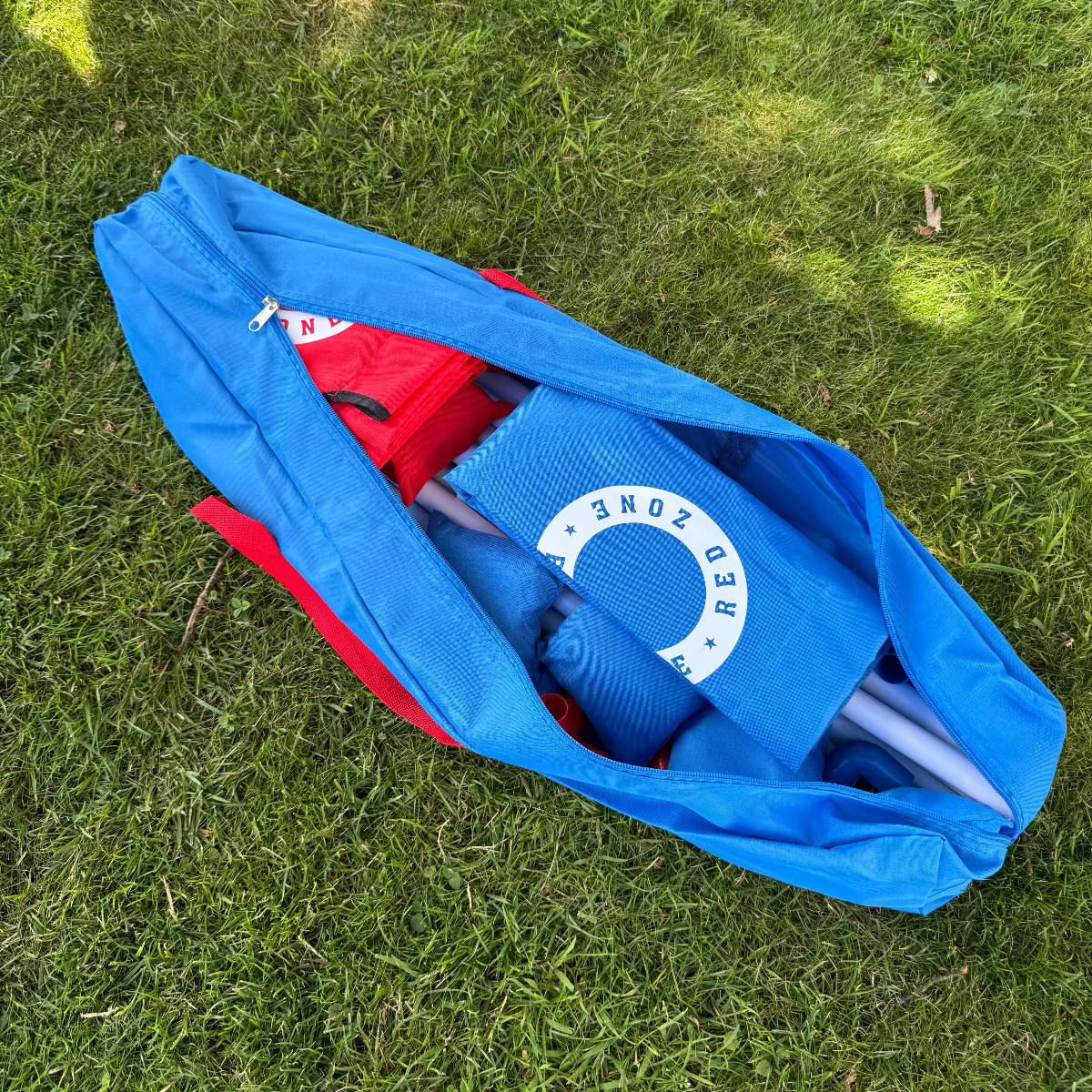 Cornhole strand sæt pakket ned i en let og smart bæretaske som medfølge på grønt græs. Tasken er blå med 2 røde cornholebræt og 8 røde og blå cornhole kasteposer, cornholeposer og ærteposer til et ærteposekastespil.