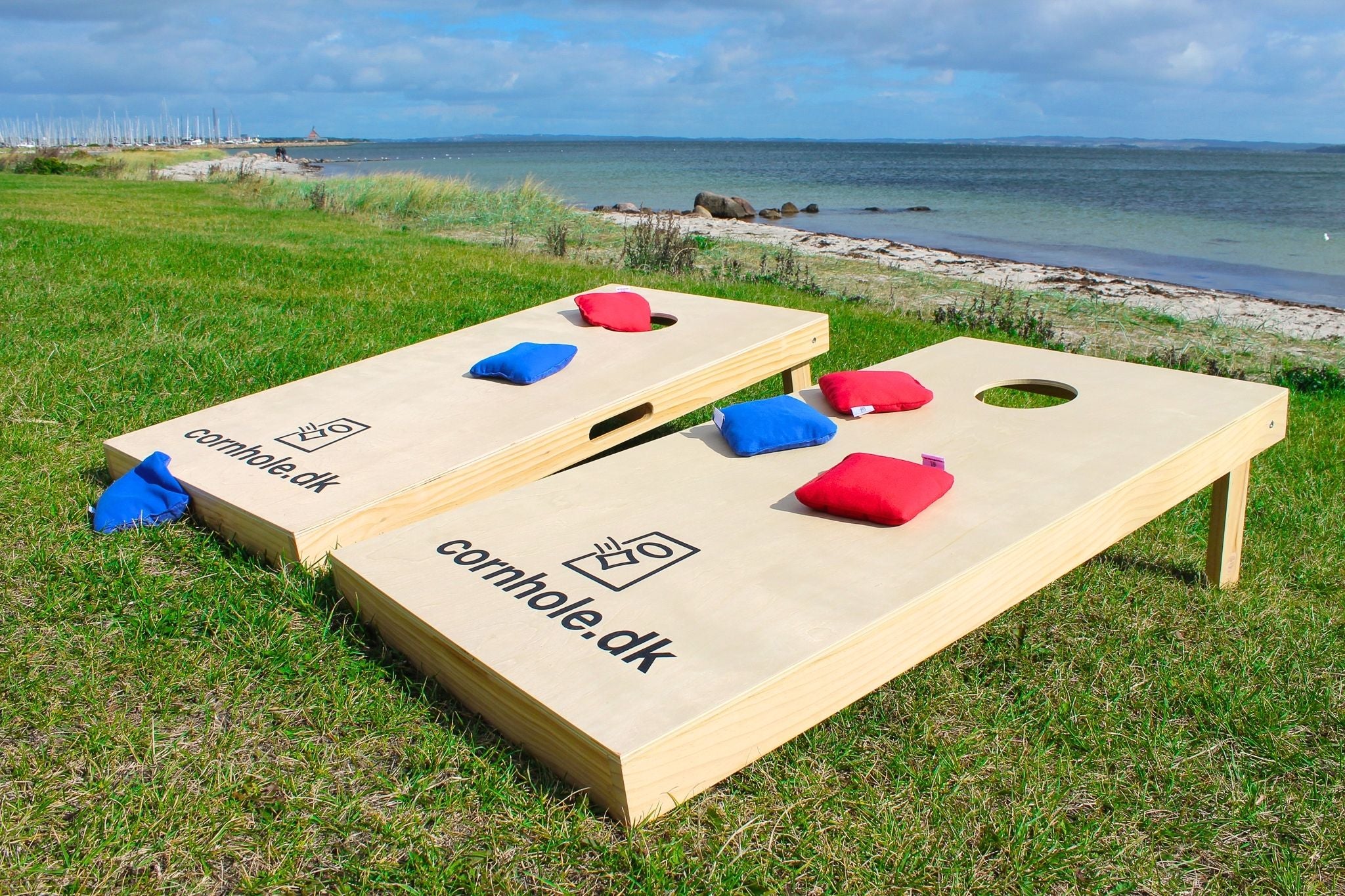 Cornholesæt i de officielle mål 120x60 cm fra cornhole.dk står på en græsplæne foran en strand med cornholeposer i farverne rød og blå fra cornhole.dk