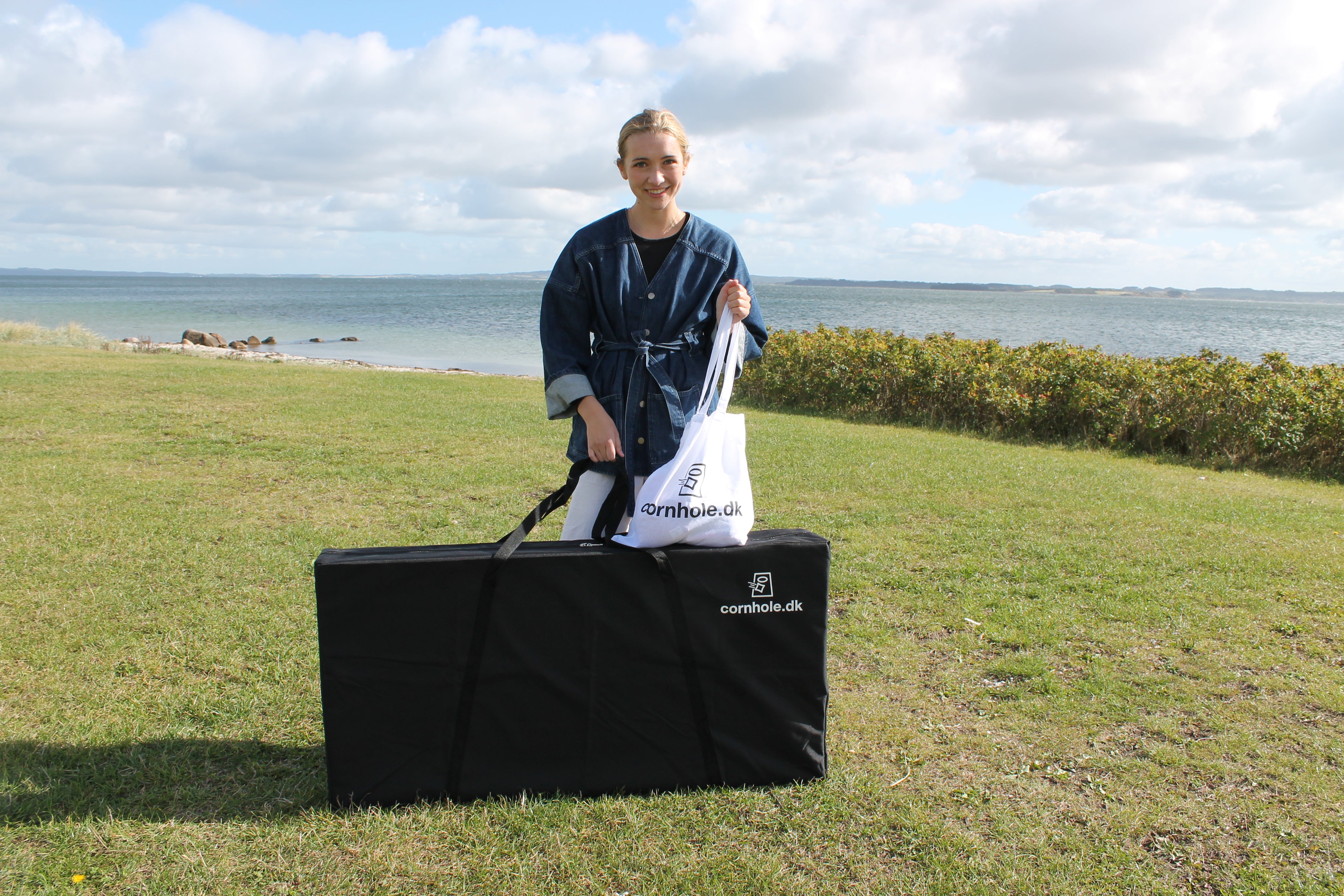 cornhole spil med pige og mulepose med cornhole.dk logo står på en græsplæne ved havet og standen